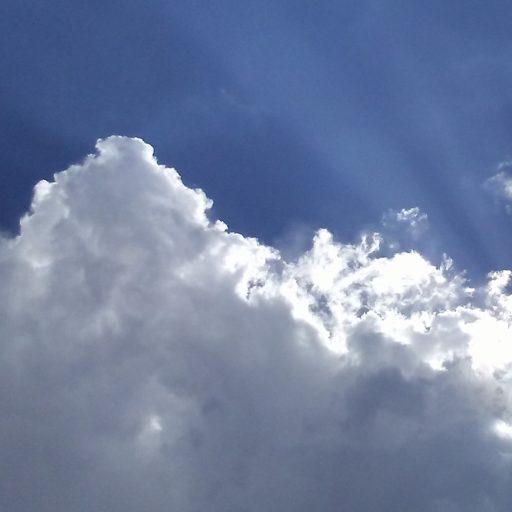 cumulus clouds, white and grey, with deep blue sky behind and rays of sun emanating up from the mid-left of the picture